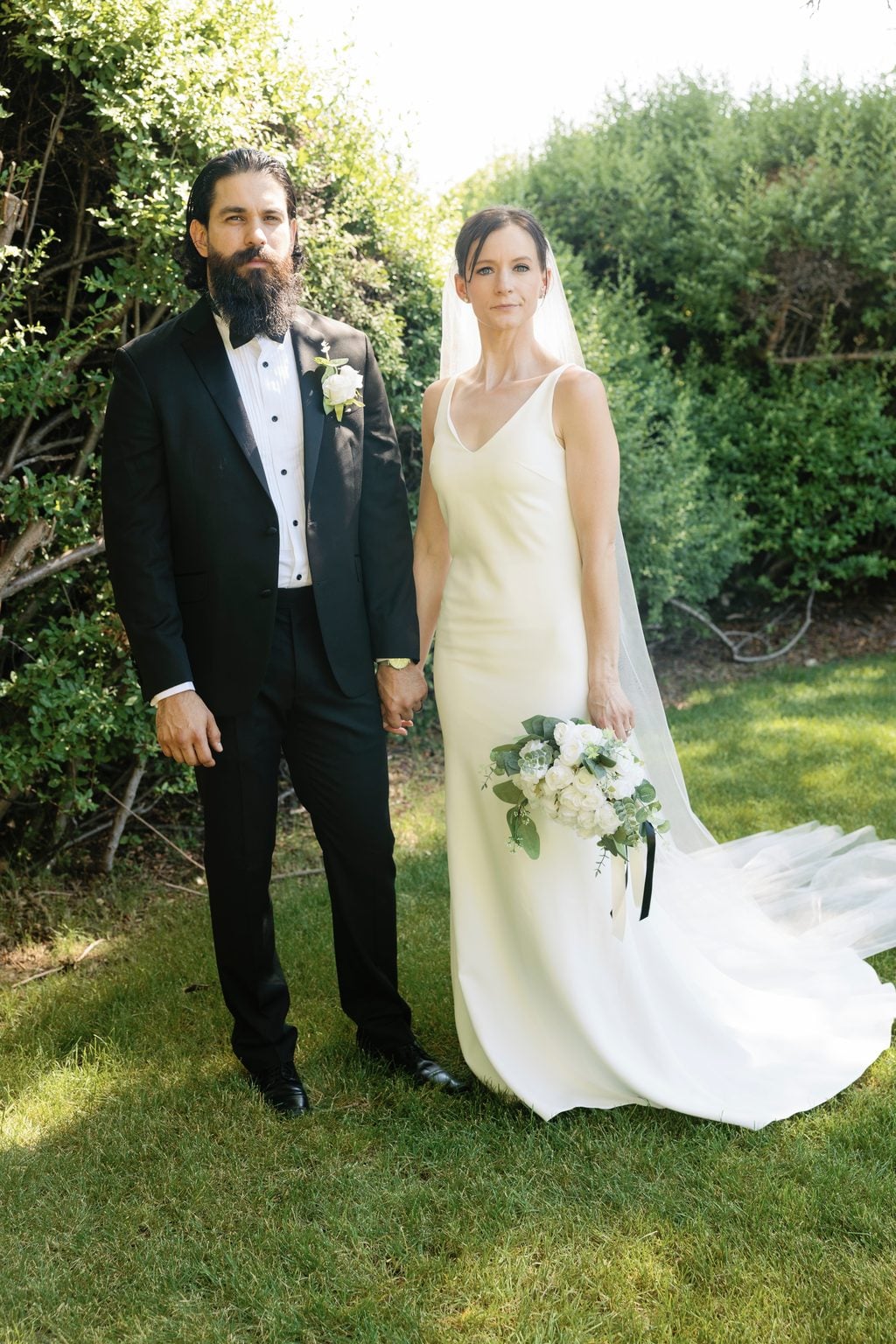 A bride in a slip gown and long veil holds her groom's hand before a garden hedge, a black-tie wedding in Utah