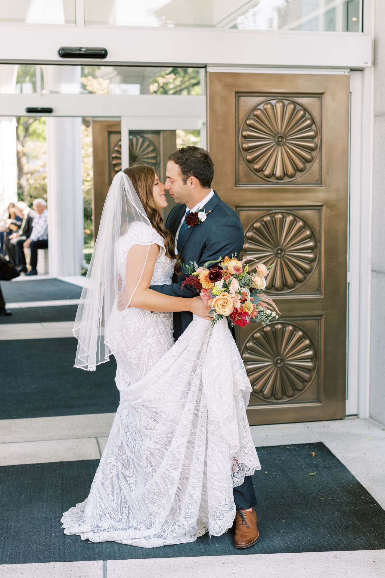 A bride and groom kiss outside ornate bronze doors just after their ceremony, with seated guests looking on