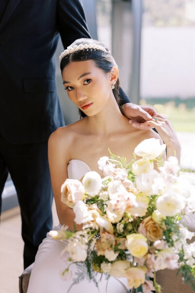 A seated bride with a pastel bouquet looks toward the camera while her groom's hand rests on her shoulder, an editorial wedding portrait in Utah