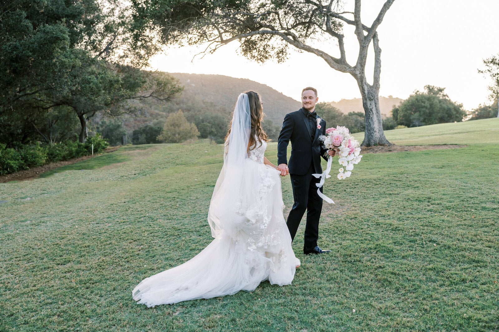 A groom holds his bride's hand on a tree-lined lawn in soft golden evening light